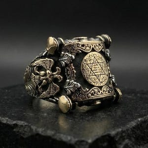 May include: An ornate ring in an antique style with a detailed design. The ring features a central emblem with a six-pointed star, surrounded by intricate patterns and embellishments. The metal has a bronze and silver finish.