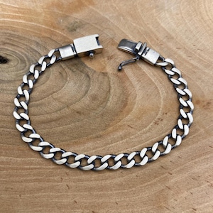 Könnte beinhalten: Silbernes Panzerkettenarmband mit sicherem Verschluss. Das Armband hat eine Reihe von ineinandergreifenden ovalen Gliedern, die ein kühnes und klassisches Design bilden. Der Verschluss ist rechteckig und hat einen sicheren Verschluss. Das Armband wird auf einer Holzoberfläche präsentiert.