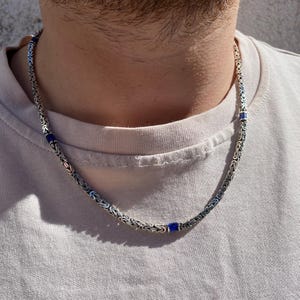 Könnte beinhalten: Silberfarbene Kette mit gewebtem Design und eingestreuten kleinen, quadratischen, dunkelblauen Akzenten. Die Halskette wird um den Hals getragen und liegt auf einem hellen T-Shirt.