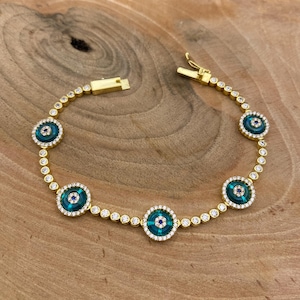 May include: Gold-toned bracelet featuring circular turquoise enamel charms with a central blue stone and a surrounding halo of small, clear crystals. The bracelet is connected by a chain of small, clear crystals.