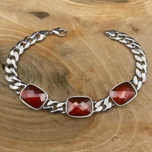 May include: A silver bracelet with a curb chain design. The bracelet features three rectangular, faceted, deep red gemstones. The clasp is a lobster claw style. The bracelet is displayed on a wooden surface.