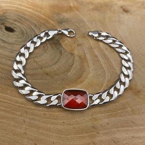 Peut inclure: Un bracelet en argent avec une chaîne gourmette. Il présente une pierre rouge rectangulaire et facettée au centre. Le bracelet est posé sur une surface en bois.