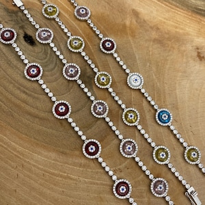 Puede incluir: Cuatro pulseras de plata con diseños circulares coloridos. Cada pulsera presenta una serie de piedras preciosas redondas engarzadas en rojo, rosa, amarillo y azul, intercaladas con pequeñas piedras transparentes. Las pulseras se exhiben sobre una superficie de madera.