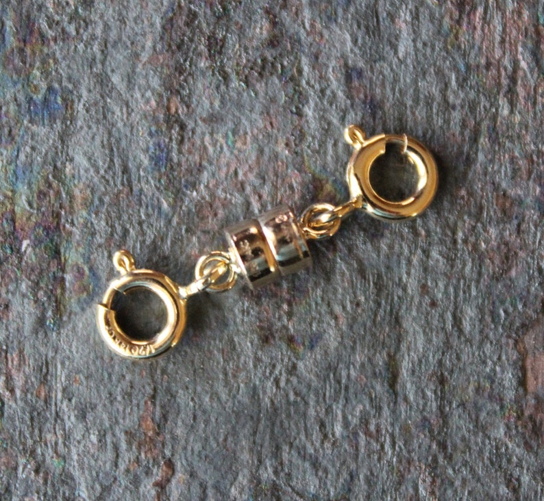 May include: A gold-colored magnetic jewelry clasp with two spring ring clasps and a cylindrical magnetic closure. The clasp is designed to connect jewelry pieces. The background is a dark, textured surface.