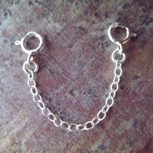 May include: A silver-colored necklace extender with two spring ring clasps connected by a delicate chain. The chain is composed of small, oval links. The extender is designed to add length to necklaces or bracelets.