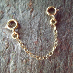 May include: A gold-colored necklace extender with two spring ring clasps connected by a delicate chain. The extender is designed to add length to necklaces or bracelets. The clasps are round and the chain is made of small, oval links.