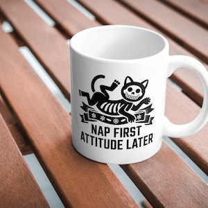 Könnte beinhalten: Weiße Keramiktasse mit einer schwarzen Grafik eines Katzen-Skeletts und dem Text "NAP FIRST ATTITUDE LATER". Die Tasse hat einen gebogenen Henkel und steht auf einer Holzoberfläche.