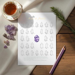 Peut inclure: Une feuille blanche avec le titre "Gems to Jewels" et des illustrations de cristaux. Un cristal violet est au centre. Une tasse de thé, des cristaux d'améthyste et deux crayons violets sont également présents.