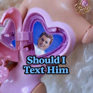 Pode incluir: Um medalhão rosa em forma de coração com uma imagem em miniatura de um homem no interior. O medalhão está aberto, revelando a imagem contra um fundo azul. O texto no medalhão diz "Should I Text Him."