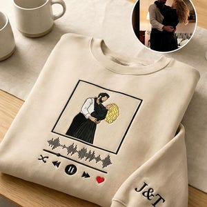 May include: Beige sweatshirt with a black and white embroidered image of a couple embracing, framed within a black square. Features a music player graphic, a red heart, and the initials "J&T" on the sleeve. Two beige mugs are in the background.