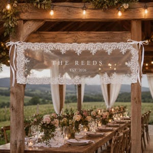 May include: A rectangular white lace banner featuring the text THE REEDS and EST. 2026 in a serif font. The banner is bordered by intricate floral lace patterns and is secured to wooden posts with white satin ribbons at each top corner.