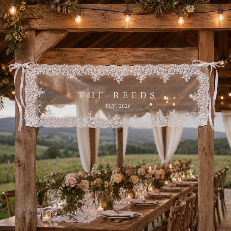 May include: A rectangular white lace banner featuring the text THE REEDS and EST. 2026 in a serif font. The banner is bordered by intricate floral lace patterns and is secured to wooden posts with white satin ribbons at each top corner.