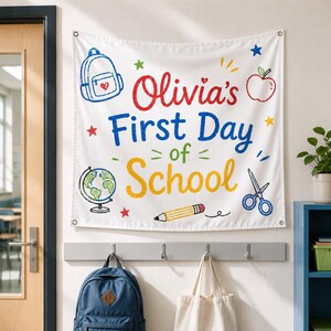 May include: A white fabric wall banner featuring the text Olivia's First Day of School in red, blue, and yellow. The design includes colourful illustrations of a backpack, an apple, a globe, a pencil, scissors, and stars printed on the fabric.