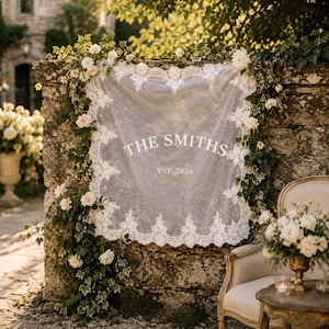 May include: A square white lace fabric sign featuring the text THE SMITHS and EST. 2026 in white lettering. The fabric has an intricate floral lace border and is draped against a stone wall adorned with green ivy vines and white roses.