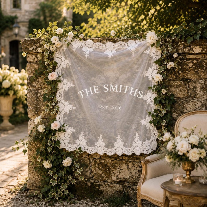 May include: A square white lace fabric sign featuring the text THE SMITHS and EST. 2026 in white lettering. The fabric has an intricate floral lace border and is draped against a stone wall adorned with green ivy vines and white roses.