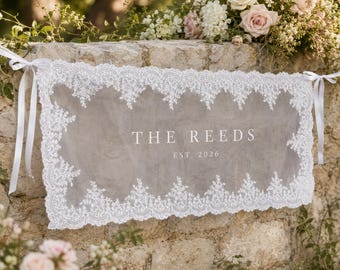 Puede incluir: Un estandarte rectangular de encaje blanco con un panel central transparente que lleva el texto impreso THE REEDS EST. 2026. El estandarte está enmarcado por un intrincado borde de encaje floral y sujeto en ambos extremos con cintas de satén blanco.