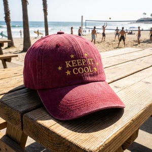 May include: A maroon baseball cap with the text "KEEP IT COOL" embroidered in gold. The hat is resting on a wooden picnic table on a sunny beach, with people playing volleyball in the background.