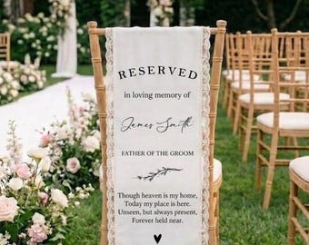 May include: A reserved sign for a wedding ceremony. The white fabric banner is draped over a wooden chair and trimmed with lace. The text reads "RESERVED" and "in loving memory of James Smith, Father of the Groom."