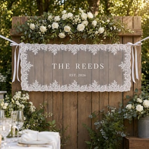 May include: A rectangular wooden sign featuring a white lace overlay with scalloped edges, tied with white ribbons. The center displays the white text THE REEDS and EST. 2026. The sign is decorated with a floral arrangement of white roses and greenery.