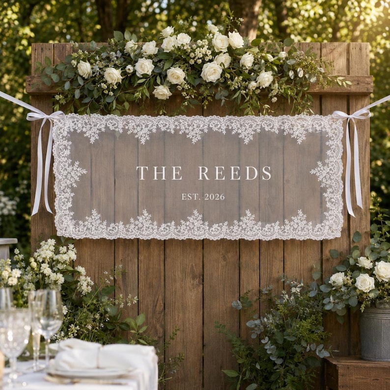 May include: A rectangular wooden sign featuring a white lace overlay with scalloped edges, tied with white ribbons. The center displays the white text THE REEDS and EST. 2026. The sign is decorated with a floral arrangement of white roses and greenery.