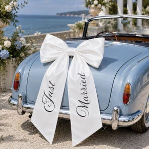 Op de afbeelding: Een grote witte satijnen strik bevestigd op de kofferbak van een lichtblauwe vintage cabriolet. De twee lange uiteinden van de strik zijn voorzien van zwarte cursieve tekst met de woorden Just Married, met Just links en Married rechts.