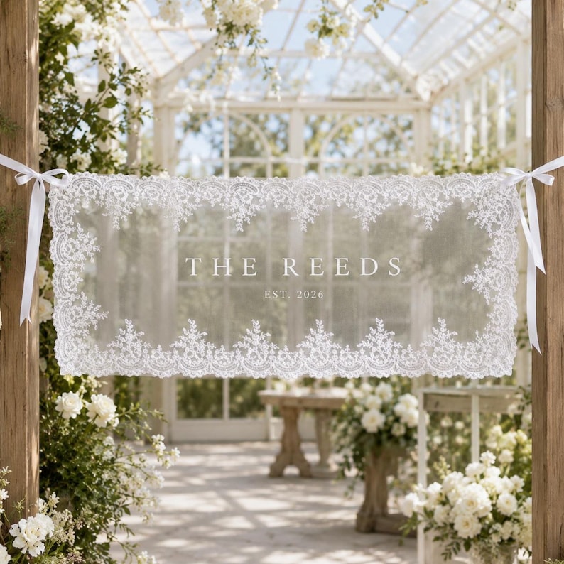 May include: A rectangular white lace banner featuring intricate floral embroidery along the edges. The center displays the text THE REEDS and EST. 2026 in a clean serif font. The banner is suspended by white ribbons tied to wooden posts.