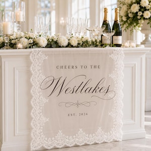 May include: A cream-colored fabric table runner with intricate white lace borders. The center features dark brown printed text that reads "CHEERS TO THE Westlakes" followed by a decorative flourish and the text "EST. 2026" in a simple serif font.