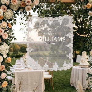 May include: A rectangular white lace-trimmed veil hanging from a wooden frame, featuring the printed text "the WILLIAMS EST.2026" in the center. The delicate fabric has an intricate floral lace border and is suspended by two gold metal clips.