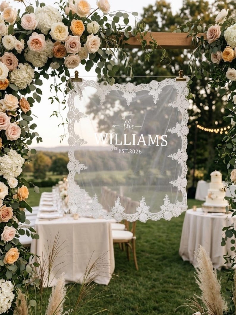 May include: A rectangular white lace-trimmed veil hanging from a wooden frame, featuring the printed text "the WILLIAMS EST.2026" in the center. The delicate fabric has an intricate floral lace border and is suspended by two gold metal clips.