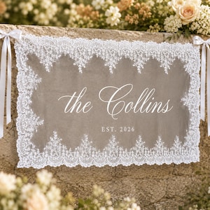 May include: A rectangular taupe fabric sign featuring a white lace border and white satin ribbons on each side. The text reads the Collins in a cursive script with EST. 2026 printed in a clean sans-serif font centered below the name.