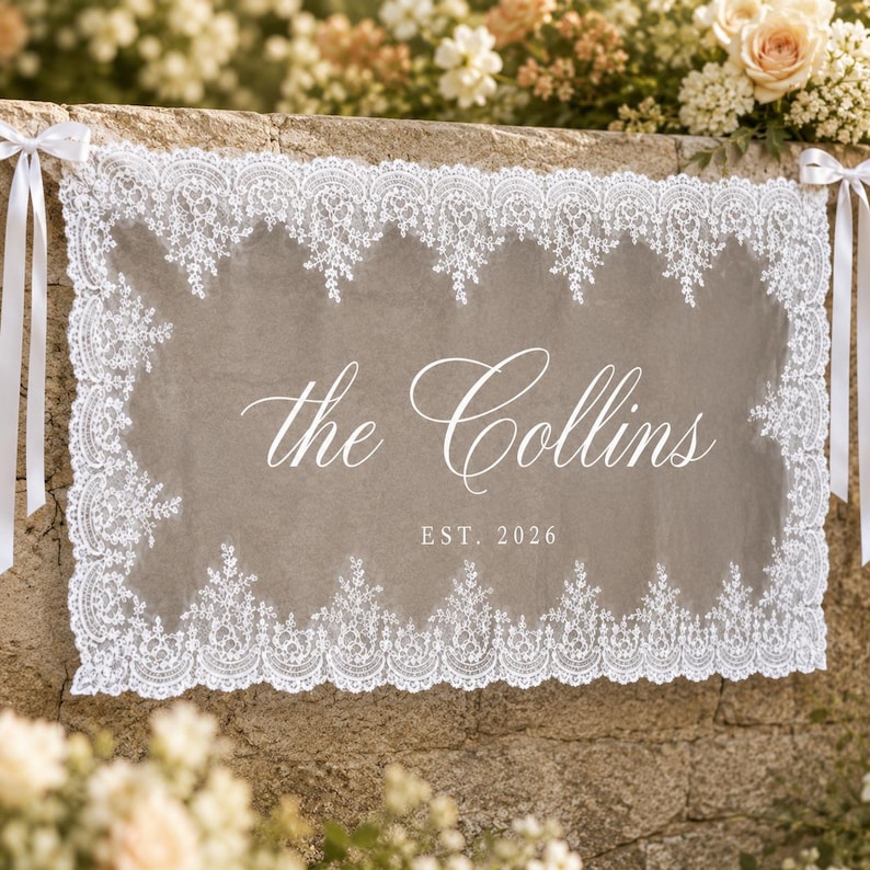 May include: A rectangular taupe fabric sign featuring a white lace border and white satin ribbons on each side. The text reads the Collins in a cursive script with EST. 2026 printed in a clean sans-serif font centered below the name.