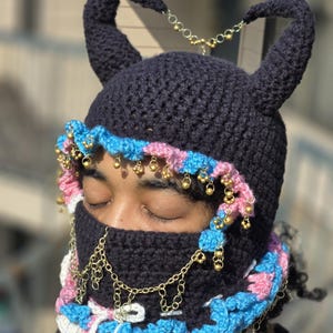 May include: A dark navy blue crochet balaclava with two horns, adorned with gold-colored chains and small bells. The balaclava features a trim of pink, light blue, and white crochet. A unique, handmade accessory.