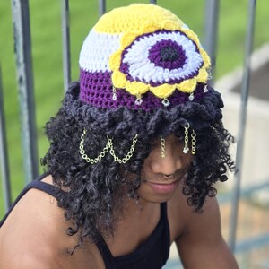 May include: A handmade crochet hat with a yellow, white, and purple design. The hat features a large flower-like embellishment and dangling decorative chains. The hat is a unique accessory.