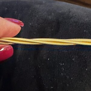 Puede incluir: Una horquilla dorada para el cabello con un diseño retorcido. La horquilla está hecha de finos hilos de metal enrollados, creando una apariencia texturizada. La horquilla se sostiene sobre un fondo oscuro.