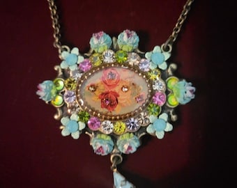 May include: A vintage-style necklace with a floral pendant. The oval pendant features a miniature painting of roses and is surrounded by blue, pink, and green crystals and flowers. A teardrop-shaped charm hangs below. The necklace is on a dark red background.