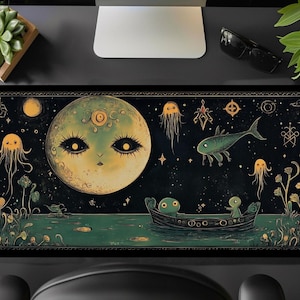 Puede incluir: Una alfombrilla de ratón verde oscuro y negro con una ilustración caprichosa de una gran luna amarilla con ojos y boca, rodeada de estrellas, peces y medusas. La luna flota sobre una masa de agua verde oscuro con un pequeño barco y dos figuras.