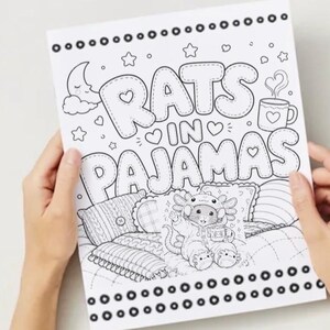 May include: A black and white coloring book page held by hands, featuring the text "RATS IN PAJAMAS". The illustration includes a cartoon rat in pajamas, pillows, a crescent moon, stars, and a mug.
