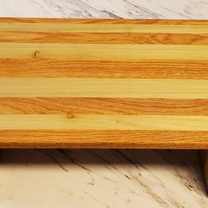 May include: A wooden riser with a natural wood finish. The top of the riser is made of alternating strips of light and dark wood. The riser has a simple design with a rectangular top and four legs.
