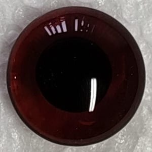 Può includere: Un occhio di vetro rotondo, rosso scuro, con una pupilla nera. L'occhio ha una finitura lucida e riflette la luce, creando riflessi bianchi brillanti. L'occhio è probabilmente per una bambola o un progetto artigianale.