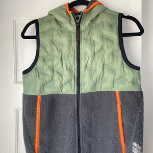 Puede incluir: Un chaleco con capucha de dos tonos. La parte superior es verde claro con un diseño acolchado, mientras que la parte inferior es de forro polar gris. Tiene detalles en naranja en la cremallera y los paneles laterales. El chaleco está colgado de una percha negra.