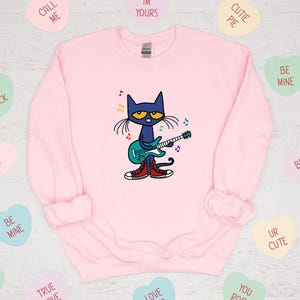 May include: Pink sweatshirt with a blue cat playing a guitar. The cat is wearing red shoes and has a blue guitar with colorful musical notes around it. The sweatshirt has a crew neck and long sleeves.