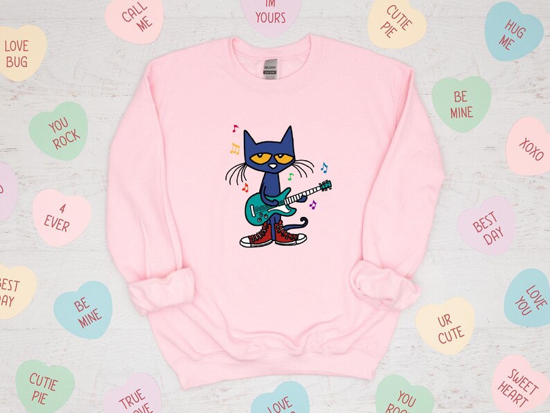 May include: Pink sweatshirt with a blue cat playing a guitar. The cat is wearing red shoes and has a blue guitar with colorful musical notes around it. The sweatshirt has a crew neck and long sleeves.