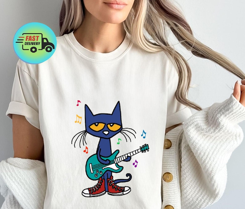 May include: A white t-shirt with a blue cat wearing red shoes and playing a green electric guitar. The cat has yellow eyes and is surrounded by musical notes.
