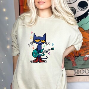 May include: A light green sweatshirt with a cartoon illustration of a blue cat playing a guitar. The cat is wearing red shoes and there are musical notes around it.