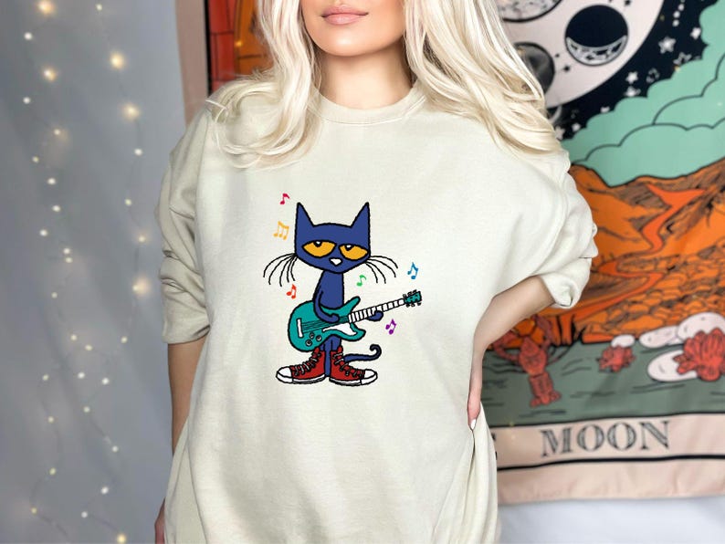 May include: A light green sweatshirt with a cartoon illustration of a blue cat playing a guitar. The cat is wearing red shoes and there are musical notes around it.