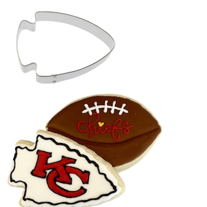 May include: Two sugar cookies decorated with icing. One cookie is shaped like a football with the word "Chiefs" written on it. The other cookie is shaped like an arrowhead with the letters "KC" in red and white inside. A metal cookie cutter shaped like a football is also shown.