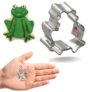 Peut inclure: Un emporte-pièce en métal argenté en forme de grenouille avec un bord ondulé. L'emporte-pièce est présenté dans une main et à côté d'un biscuit en forme de grenouille verte.