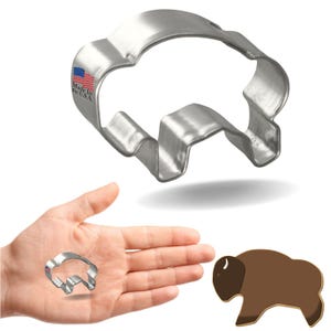 May include: A silver metal cookie cutter shaped like a bison. The cookie cutter is shown on a white background with a hand holding a smaller version of the cookie cutter. A brown cookie shaped like a bison is also shown.
