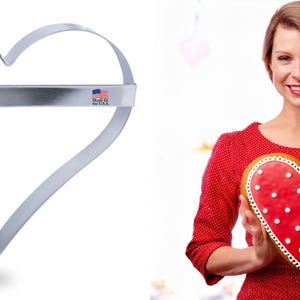 May include: A silver heart-shaped cookie cutter with a metal bar across the top. The cutter is made in the USA. A woman is holding a red heart-shaped cookie with white polka dots.