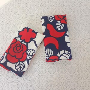 May include: Two folded cloth napkins with a floral pattern. The design features large red roses and white leaves on a navy blue background. The napkins are on a white lace surface.